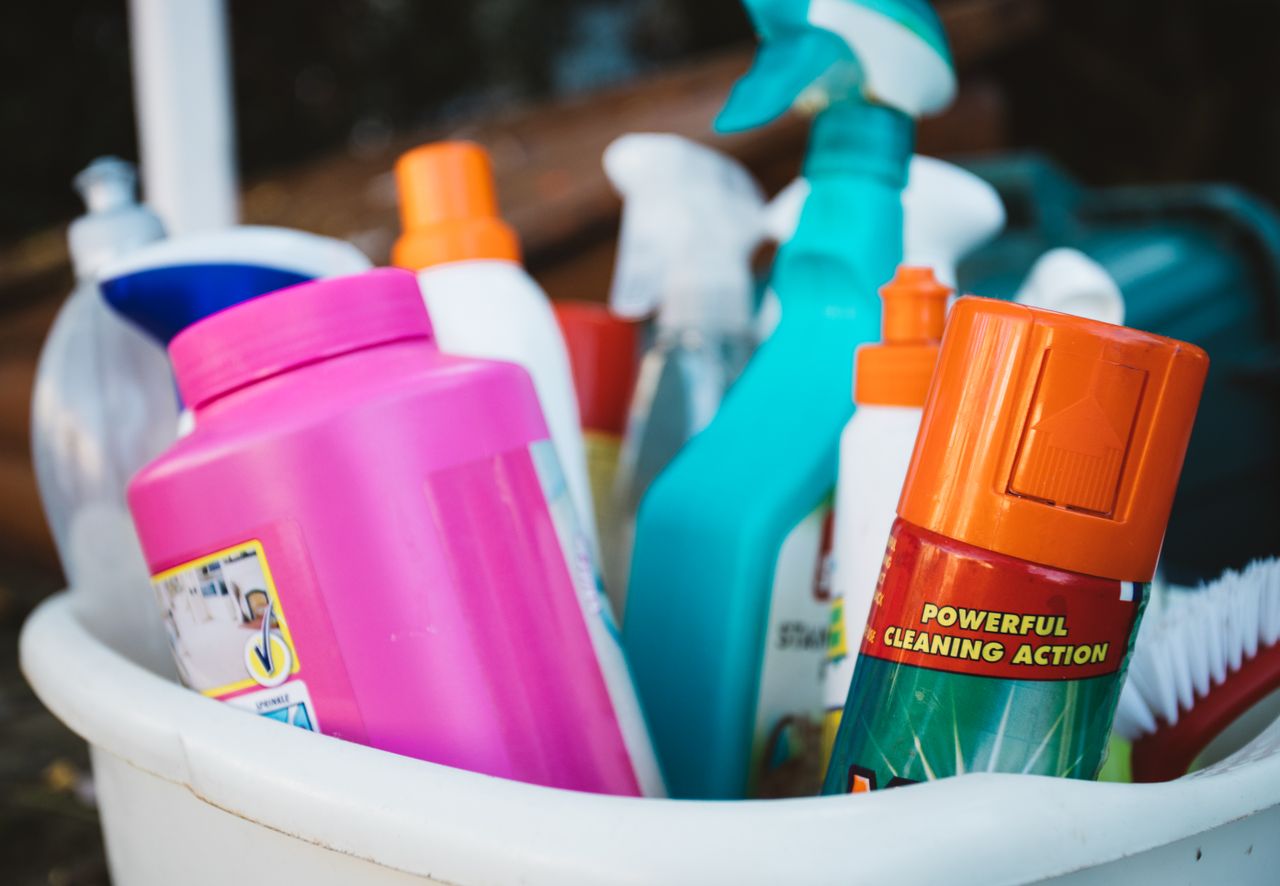 Gros plan sur divers produits de nettoyage rassemblés dans un seau en extérieur, mettant en avant différents détergents et sprays.